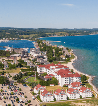 Aerial view of a scenic Northern Michigan lakeside town with historic red-roofed buildings, waterfront homes, and a marina along clear blue waters, showcasing local charm and inviting outdoor adventures.