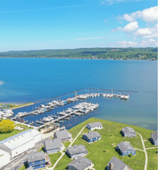 Aerial view of a marina with boats docked along piers on a clear blue lake, surrounded by green lawns and small cottages, showcasing a serene Northern Michigan waterfront community ideal for local adventures and hidden gem exploration.