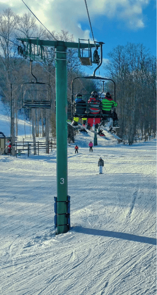 Skiers riding a chairlift over a snowy slope surrounded by bare trees under a clear blue sky in Northern Michigan, showcasing winter outdoor adventures and local activities.
