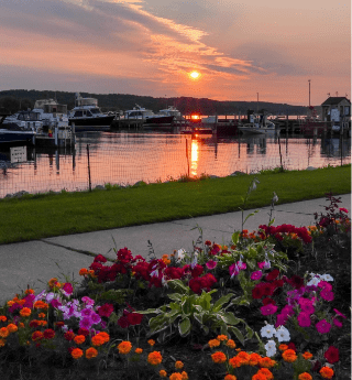 Sunset over a marina in Northern Michigan with boats docked on calm water, colorful flowerbeds in the foreground, and a scenic waterfront walkway, showcasing local hidden gems and outdoor adventures.