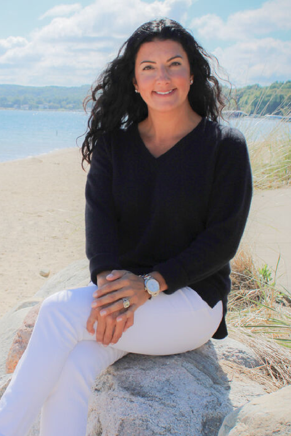 Image 1 Woman with long dark curly hair wearing a black sweater and white pants sitting on a rock by a sandy beach with water and trees in the background, representing a Northern Michigan local and real estate expert sharing curated recommendations for exploring hidden gems and adventures in the region.