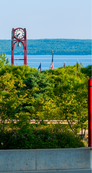 Red clock tower and American flags overlooking lush green trees with Lake Michigan and forested hills in the background, representing a scenic Northern Michigan local attraction and outdoor adventure destination.