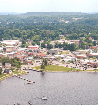 Aerial view of a Northern Michigan lakeside town featuring a marina, waterfront park, and a mix of downtown buildings surrounded by lush green forested hills.